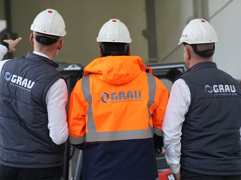 Three GRAU staff in hard hats and branded workwear, seen from behind on the factory floor