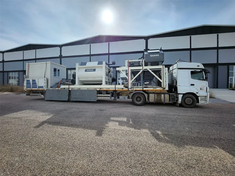Composants de la centrale à béton fixe GRAU G135S chargés sur un camion plateau devant un hall d’usine