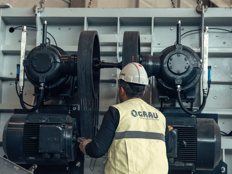 Ouvrier en gilet GRAU et casque, vu de dos, travaillant sur les deux moteurs et transmissions par courroie d’une machine
