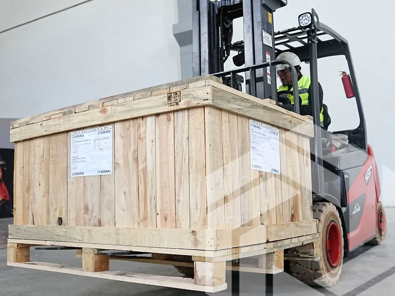 Crated GRAU spare parts lifted by a forklift indoors, the driver in a hard hat and high-visibility vest